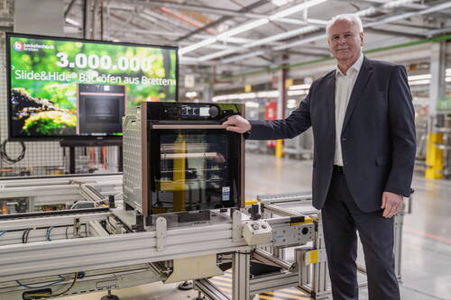 Geschäftsmann in Anzug präsentiert auf dem Förderband einer Fabrik einen Slide&Hide-Backofen, während im Hintergrund ein großer Bildschirm die Produktion des 3.000.000sten Geräts in Bretten verkündet.