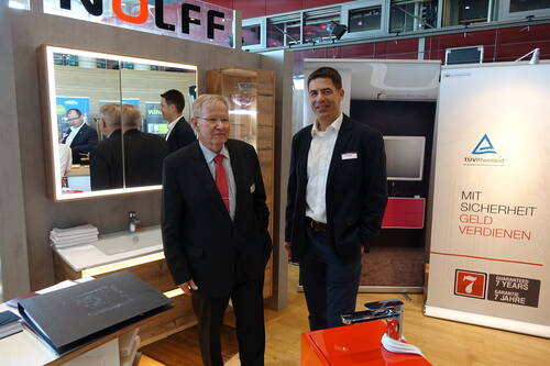 Zwei Geschäftsleute im Anzug stehen an einem Messestand vor einem beleuchteten Badspiegel und einem TÜV-Rheinland-Roll-up mit der Aufschrift „Mit Sicherheit Geld verdienen“.