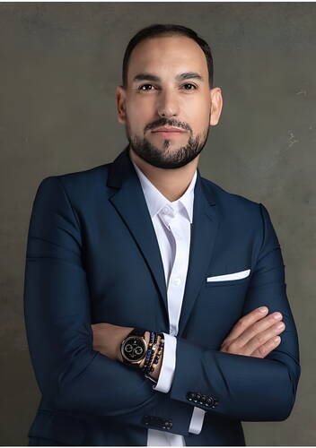 Selbstbewusst dreinblickender Mann im dunkelblauen Anzug mit weißem Hemd, verschränkten Armen und eleganter Armbanduhr vor neutralem Hintergrund