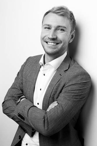 Lächelnder junger Mann im Anzug mit verschränkten Armen lehnt entspannt an einer weißen Wand in einem schwarz-weißen Porträt