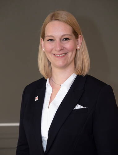 Lächelnde blonde Frau im schwarzen Blazer und weißer Bluse posiert vor einfarbigem Hintergrund in professioneller Porträtaufnahme.