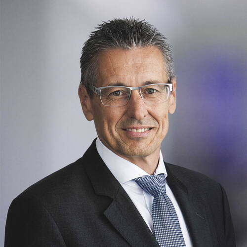Älterer Geschäftsmann mit grauen Haaren, Brille und Anzug lächelt selbstbewusst vor neutralem Hintergrund.
