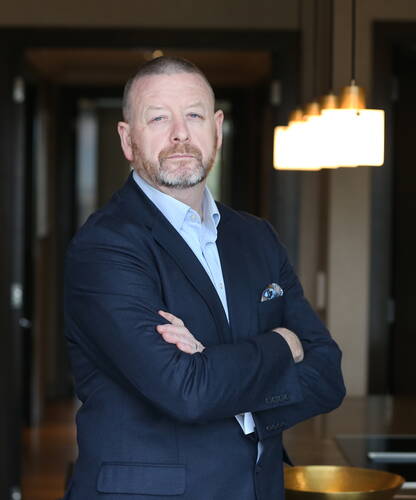 Selbstbewusster Mann mittleren Alters im Businessanzug steht mit verschränkten Armen in einem modern beleuchteten Büroflur
