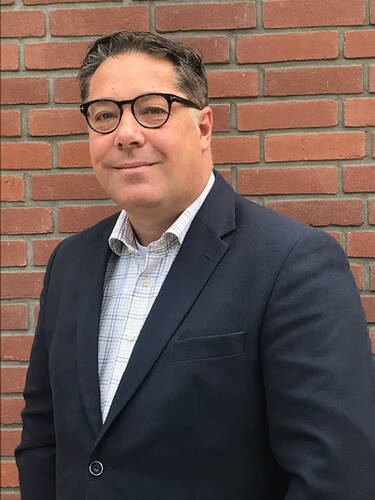 Mittelalterlicher Mann mit Brille im dunklen Sakko und kariertem Hemd steht lächelnd vor einer roten Backsteinwand