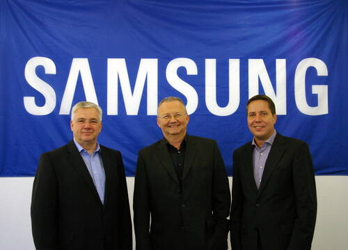 Drei lächelnde Männer in Anzügen posieren vor einem großen blauen Banner mit dem weißen Samsung-Schriftzug