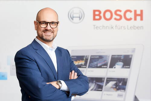 Geschäftsmann im blauen Anzug steht lächelnd mit verschränkten Armen vor einem Bosch‑Werbehintergrund mit dem Slogan „Technik fürs Leben“ und einem großen Bildschirm mit Produktübersicht