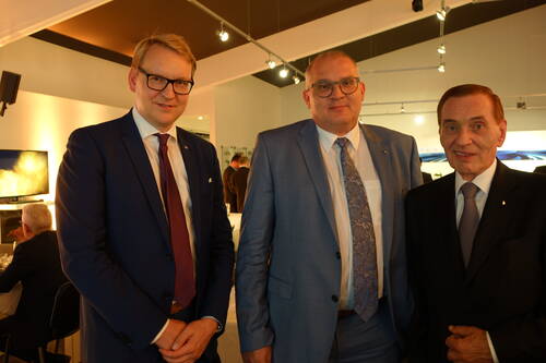 Drei elegant gekleidete Männer in Anzügen stehen nebeneinander und lächeln in die Kamera, im Hintergrund ein hell beleuchteter Veranstaltungsraum mit weiteren Gästen.
