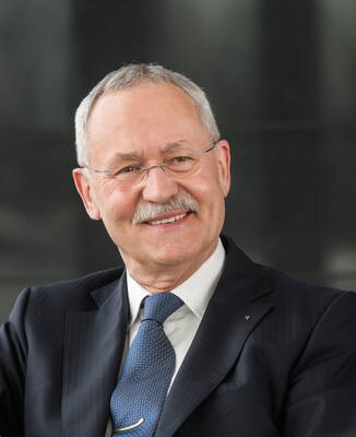 Älterer Mann im Anzug mit Krawatte und Brille lächelt freundlich in einer professionellen Porträtaufnahme vor dunklem Hintergrund
