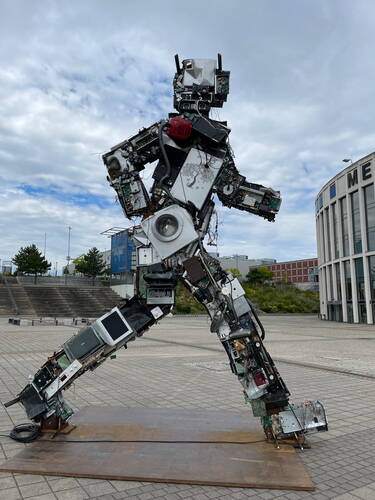 Große humanoide Skulptur aus alten Elektrogeräten in dynamischer Laufpose auf einem modernen Platz vor einem runden Messegebäude unter wolkigem Himmel