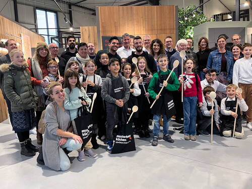 Große fröhliche Gruppe aus Kindern mit Kochschürzen und Kochlöffeln sowie Erwachsenen dahinter, die gemeinsam in einer modernen Küchenausstellung posieren.