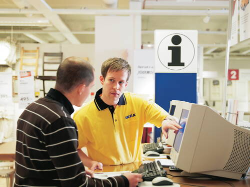 IKEA-Mitarbeiter in gelbem Polohem d erklärt einem Kunden am Computer die Produktdetails an einem Informationsschalter im Möbelhaus