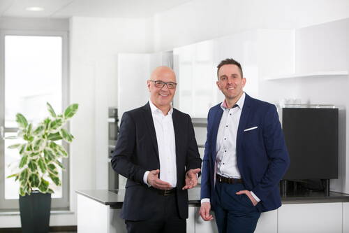 Zwei lächelnde Geschäftsmänner im Anzug stehen in einer modernen, hellen Büroküche und blicken in die Kamera