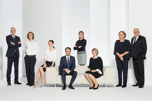Ein gemischtes Team aus acht Geschäftsleuten in formeller Kleidung posiert in einem hellen, minimalistischen Studio, wobei einige auf einem modernen Sofa sitzen und andere selbstbewusst dahinter stehen.