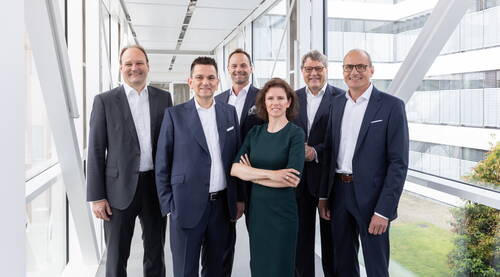 Sechs Führungskräfte in Businesskleidung posieren lächelnd in einem modernen verglasten Büroflur, die Frau in der Mitte steht mit verschränkten Armen vor der Gruppe.