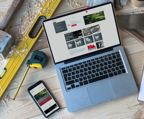 Offene Werkbank mit Laptop und Smartphone, auf denen eine Häfele-Webseite angezeigt wird, umgeben von Maßband, Wasserwaage, Säge und Holzspänen