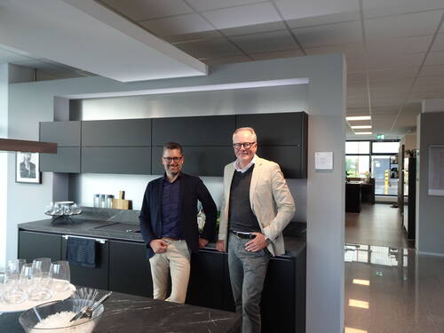 Zwei lächelnde Männer im Business-Outfit stehen locker an einer modernen schwarzen Küchenzeile in einem hellen Showroom mit Glaswaren und Küchenaccessoires auf der Arbeitsplatte