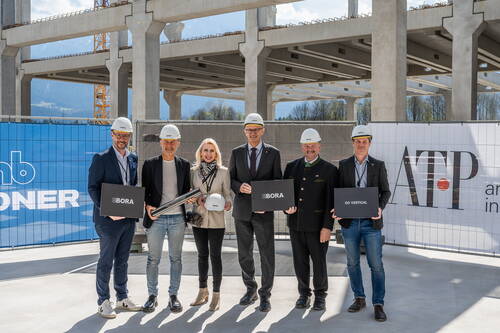 Gruppe von sechs Personen mit Schutzhelmen posiert bei sonnigem Wetter auf einer Baustelle eines modernen Industriegebäudes und hält schwarze Schilder mit den Aufschriften „BORA“ und „GO VERTICAL“ sowie eine Metallrolle in den Händen