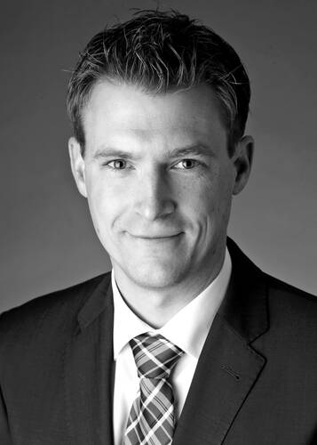 Junger Mann im Anzug mit kariertem Schlips blickt lächelnd in die Kamera vor neutralem Hintergrund