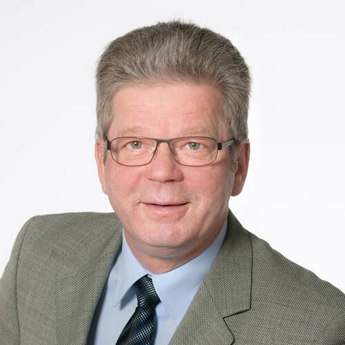 Älterer Mann mit Brille und grauen Haaren im grauen Anzug und hellblauen Hemd lächelt freundlich vor weißem Hintergrund