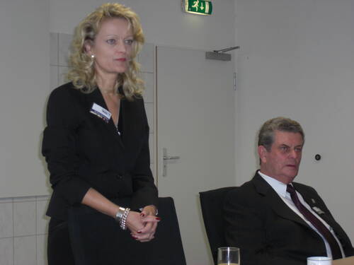 Geschäftsfrau steht im Konferenzraum und spricht, während ein älterer Mann im Anzug aufmerksam auf einem Stuhl sitzt.