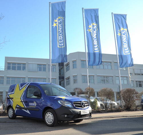Blauer Mercedes-Lieferwagen mit Euronics-Werbung steht vor einem modernen Bürogebäude mit drei Euronics-Fahnen im Wind.