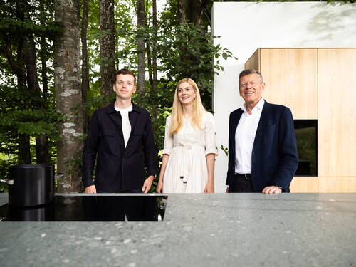 Drei Menschen stehen lächelnd hinter einer modernen Outdoor-Küche mit Induktionskochfeld, eingerahmt von Bäumen und einer hellen Holzwand im Hintergrund.