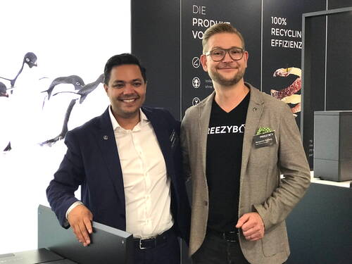Zwei lächelnde Männer in Business-Kleidung posieren auf einer Messe vor einem Stand mit Recycling-Thema und einem hell beleuchteten Pinguin-Motiv im Hintergrund.