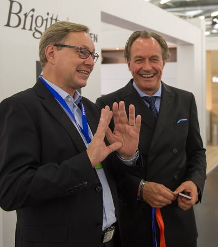 Zwei Geschäftsleute im Anzug unterhalten sich lachend auf einer Messe, während der Mann links gestikulierend etwas erklärt.