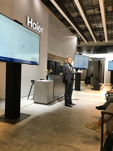 Geschäftsmann im Anzug hält vor sitzendem Publikum eine Präsentation über IoT in einem modernen Haier-Messestand mit großen Bildschirmen und minimalistischem Podium