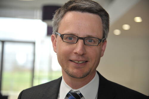 Mittelalterlicher Mann mit Brille und kurzem Bart lächelt im Business-Anzug in einem modernen, hellen Bürogebäude