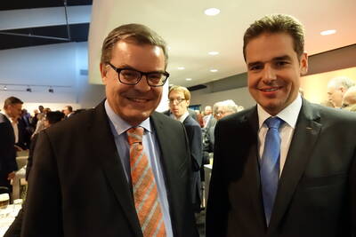 Zwei lächelnde Männer im Anzug stehen dicht beieinander bei einer formellen Netzwerkveranstaltung in einem gut besuchten Konferenzraum.