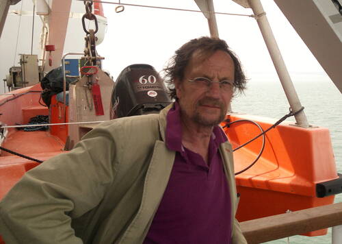 Mittelalterlicher Mann mit Brille lehnt an der Reling eines Schiffs vor einem orangefarbenen Rettungsboot auf ruhiger See