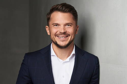 Lächelnder Mann im dunklen Anzug und weißem Hemd lehnt an einer hellgrauen Betonwand und blickt direkt in die Kamera.