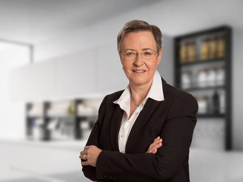Selbstbewusste Geschäftsfrau mittleren Alters mit Kurzhaarfrisur, Brille und dunklem Blazer verschränkt die Arme und lächelt in einer modernen, hellen Büroumgebung