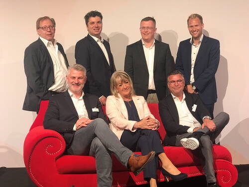 Geschäftsleute in Anzug und Blazer posieren lächelnd als Gruppe, teils stehend, teils sitzend auf einem auffälligen roten Sofa vor neutralem Hintergrund