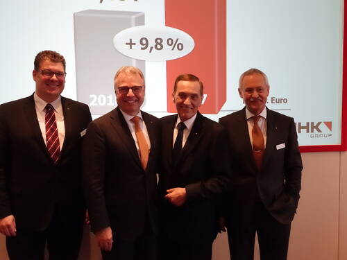 Vier Geschäftsmänner im Anzug lächeln vor einer Präsentationsfolie mit Balkendiagramm und der Kennzahl „+9,8 %“