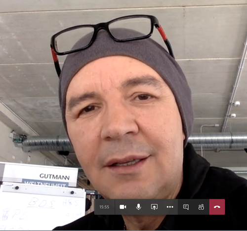 Mann mit grauer Mütze und hochgeschobener Brille spricht in einer Videokonferenz in einem modernen Industrie-Büro.