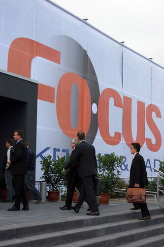 Geschäftsleute im Anzug gehen mit Aktentaschen die Eingangsstufen zu einem Konferenzgebäude mit großem „Focus“-Werbebanner hinauf