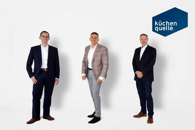 Drei Geschäftsmänner in Anzügen stehen vor weißem Hintergrund neben dem Logo des Küchenunternehmens „küchen quelle“