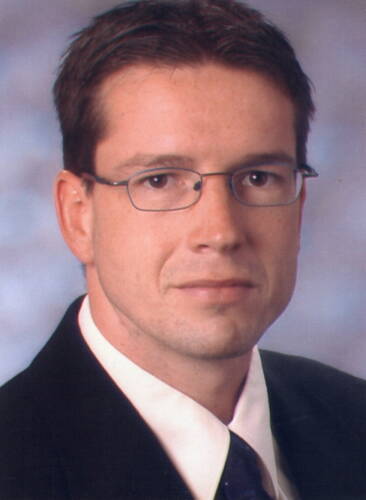 Junger Mann mit kurz geschnittenen dunklen Haaren, Brille und Anzug schaut ernst in die Kamera vor neutralem Hintergrund