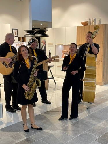 Musikgruppe in schwarzer Kleidung mit gelben Schals spielt Saxofon, Gitarre und Kontrabass in einer modern eingerichteten Küche, während eine Sängerin in der Mitte performt