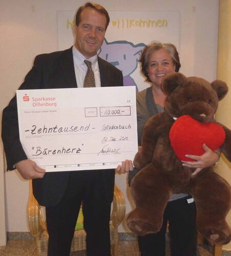 Mann im Anzug überreicht gemeinsam mit einer lächelnden Frau einen übergroßen Spendenscheck über 10.000 Euro an die Organisation „Bärenherz“, während sie einen riesigen Teddybären mit rotem Herz im Arm hält