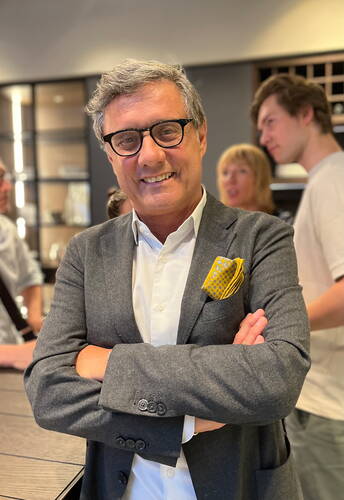 Älterer Mann mit Brille und grauem Sakko steht mit verschränkten Armen lächelnd in einem modernen, belebten Innenraum
