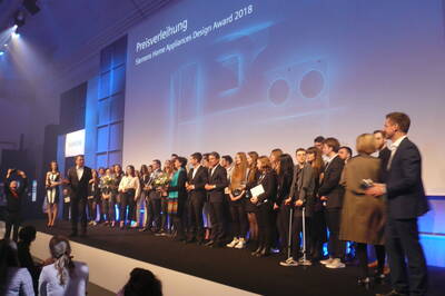 Große Gruppe junger Designerinnen und Designer steht bei der Preisverleihung des Siemens Home Appliances Design Award 2018 auf einer Bühne und erhält Auszeichnungen vor einer beleuchteten Leinwand.