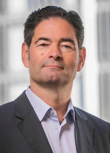 Mittelalterlicher Mann im Anzug blickt ernst und selbstbewusst in die Kamera, unscharfer Bürogebäudehintergrund im Freien.