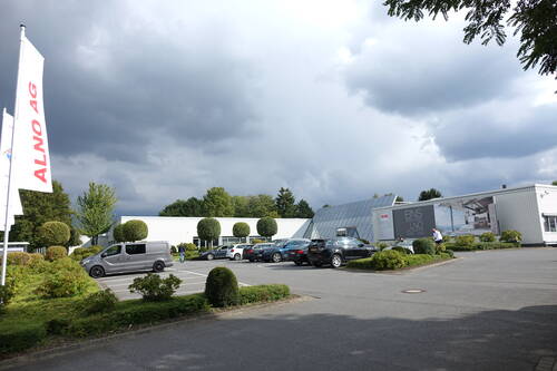 Parkplatz vor dem modernen ALNO-Firmengelände mit geparkten Autos, gepflegten Büschen und dunklen Gewitterwolken am Himmel