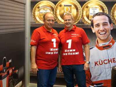 Zwei Mitarbeiter in roten „Komm ins Team“-Poloshirts stehen lächelnd vor einer Messestandwand mit goldenen Auszeichnungen und einem großen Poster eines Rennfahrers.