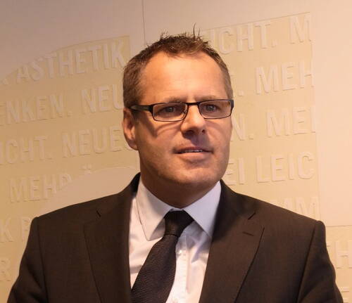 Mittelalterlicher Mann im Anzug mit Brille blickt leicht zur Seite vor einer beige beschrifteten Wand