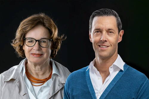 Ältere Frau mit Brille und jüngerer Mann in blauem Pullover stehen nebeneinander vor dunklem Hintergrund und lächeln in die Kamera.