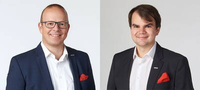 Zwei lächelnde Männer in dunklen Anzügen mit weißen Hemden und roten Einstecktüchern stehen vor hellem Hintergrund nebeneinander in einem professionellen Porträt.